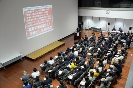 大型のスクリーンを前に座って講演を聴く聴衆を斜め上から撮影した写真