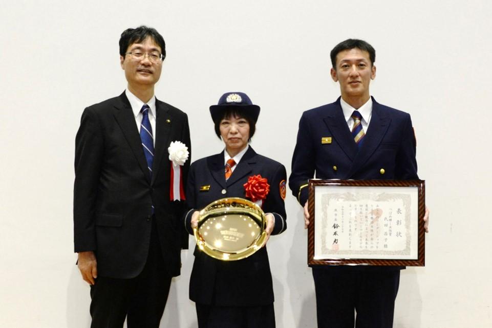 両側に男性、真ん中に女性が楯を持って表彰されている写真