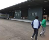 突き出た屋根のある灰色の建物の入り口付近を二人の人物が歩いている写真