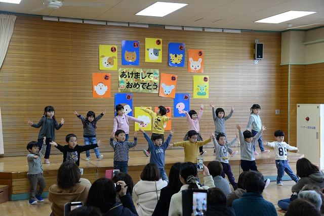 ステージで歌と踊りを披露する子どもたちとそれを見守る保護者たちの写真
