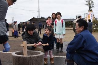 臼と杵を使い、餅つき体験をしている女の子と保護者の写真