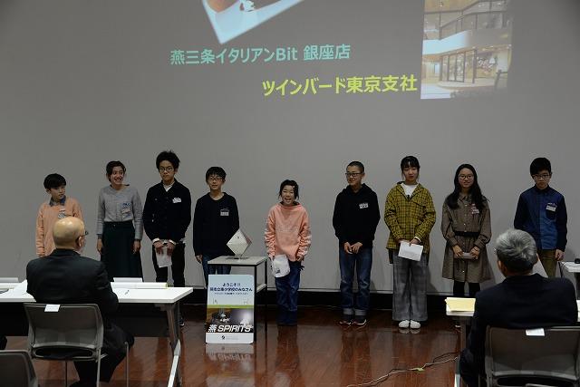 スクリーンを後ろに横に並んで立っている私服の少年少女達9人と、それを着席して見ている参加者の写真