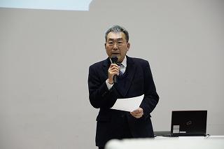 書類とマイクを手に、話をしている白髪混じりの男性の写真