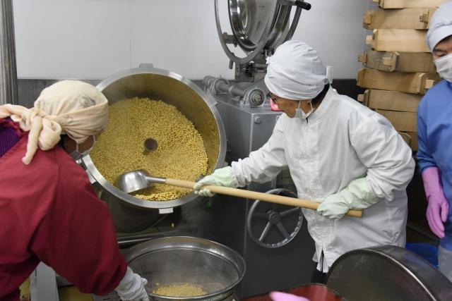 たくさんの大豆が入った圧力釜を傾け、器具を使って別の容れ物に移している女性達の写真
