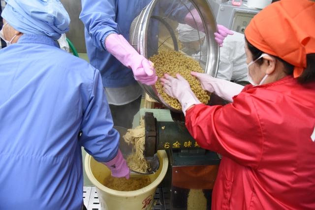 女性達がボウルの中の大豆を機械に入れ、マッシュされた状態の大豆が機械の横から出てきている様子を収めた写真