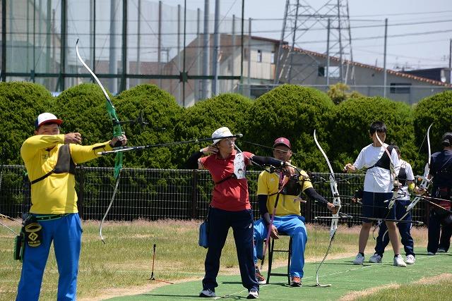 屋外で横に並びアーチェリーの弓を構えている男性と子ども数人の写真