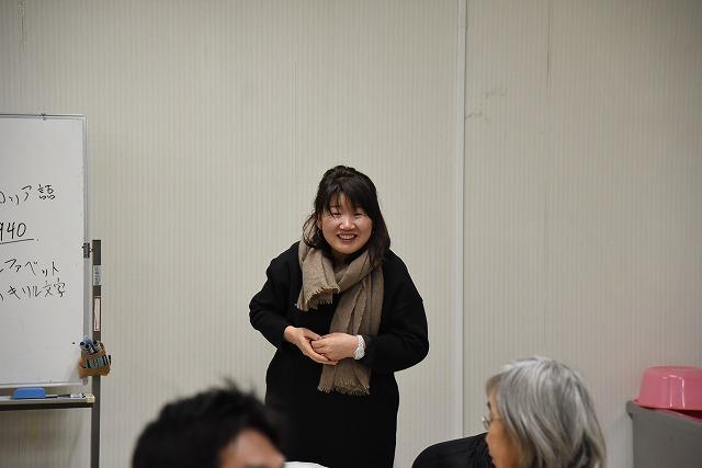 ホワイトボードを横に、笑顔で話をしているマフラーをした女性の写真
