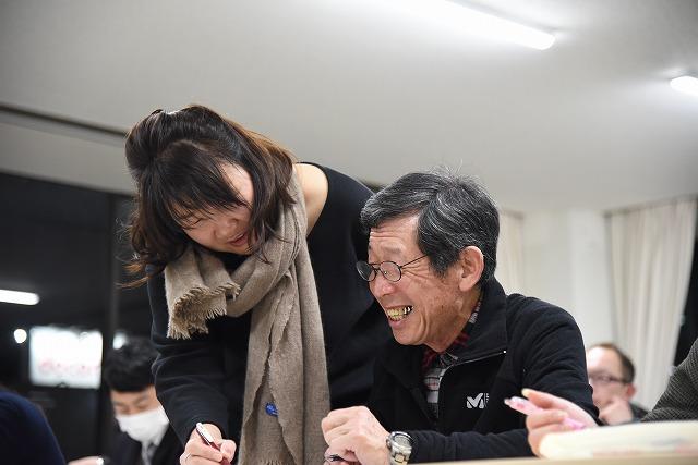 着席している参加者の中年男性の横に立ち、楽しそうに話をしながらペンで男性のプリントに何かを書き込んでいる女性の写真