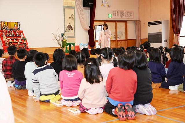 広い大きな部屋の中、ひな壇と掛け軸を横に、大勢の子ども達が座っている前で、マイクを手に話をしている女性の写真