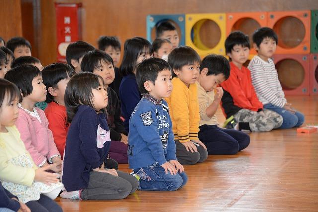 木張りの部屋の床に座って、前を向いて話を聞いているたくさんの子ども達の写真