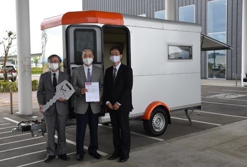 銀色のトレーラーの前に並んでいる三人の男性の集合写真