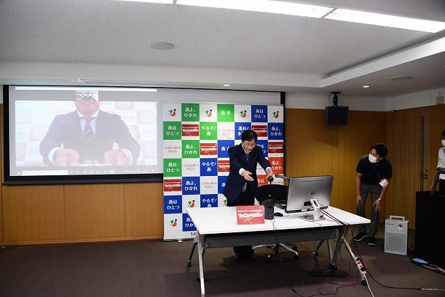 男性の写っているスクリーンの隣で、燕市のボードの前に立ち、両手を受け取るように差し出している市長の写真