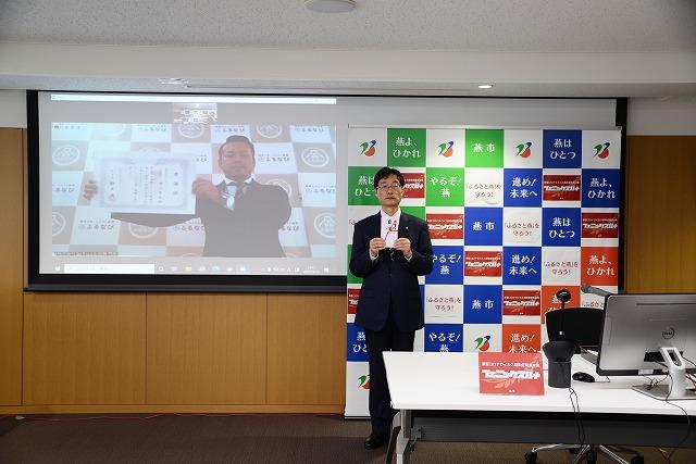 封筒を持って立っている市長の横で、スクリーンの中から感謝状を持ってこちらに見せている男性の写真