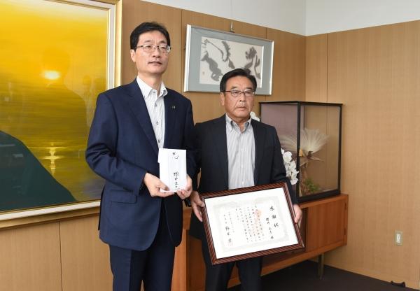 市役所の一室で、感謝状を持った男性と、白い封筒を持った男性が並んで立っている写真