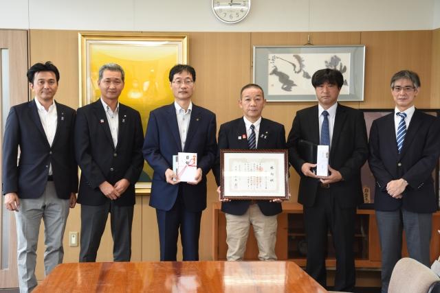6人の男性が並び、感謝状と目録などをカメラに向けている集合写真
