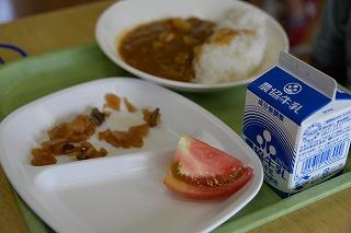 給食のトレーにのせられた牛乳パックとお皿に盛り付けられたカレーライスにトマトと福神漬けの写真