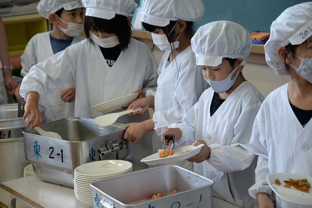 給食の割烹着を着てマスクを付け、給食を盛り付けている子供たちの写真
