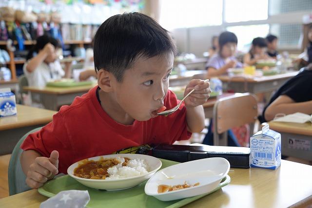 机に座って給食のトマトをスプーンですくって食べている赤いシャツを着た男の子の写真