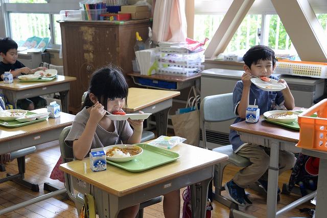 それぞれ机に座って給食のトマトをスプーンですくって食べる男の子と女の子の写真