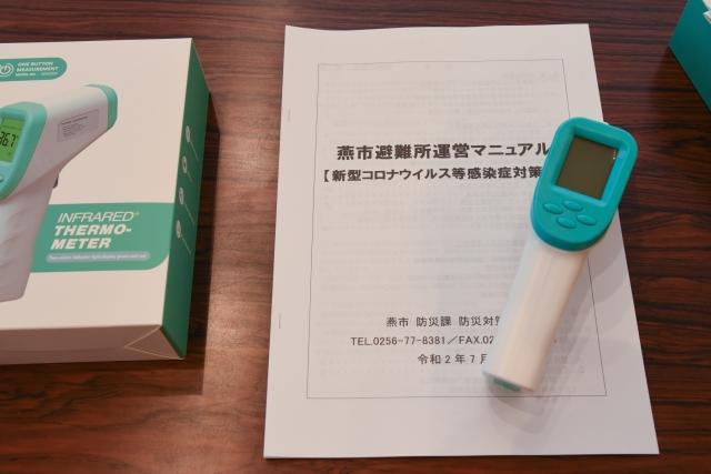 机の上に置かれた燕市避難所運営マニュアルと書かれた冊子と、ハンドタイプの非接触型体温計の写真
