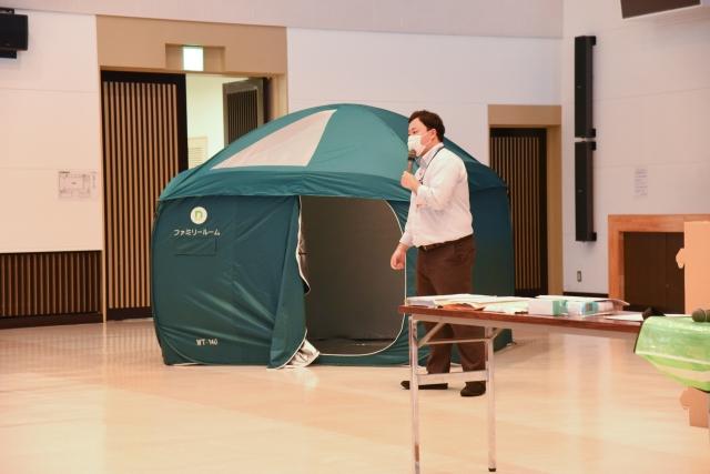 屋内に張られた避難用テントを横に、マスクを付けマイクを手に解説する防災担当職員の写真