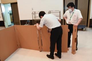 屋内で段ボール製のパーティションを組み立てているマスクを付けた2人の職員の写真