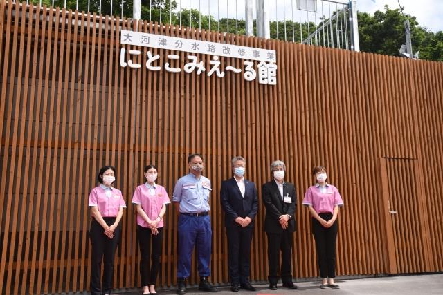 茶色い細い建材を縦にびっしり並べた「えこみえ~る館」の壁の前でそこに取り付けられた看板の下に並ぶ、マスクを付けた6人の男女の職員たちの写真