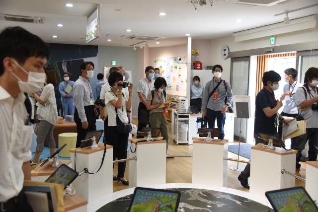 館内の展示室で円卓上に並ぶタブレットと、その様子を撮影している大勢の報道陣たちの写真
