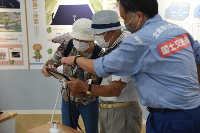 展示室内で実際にタブレットを操作する2人の来館者と隣に立って説明している男性職員の写真