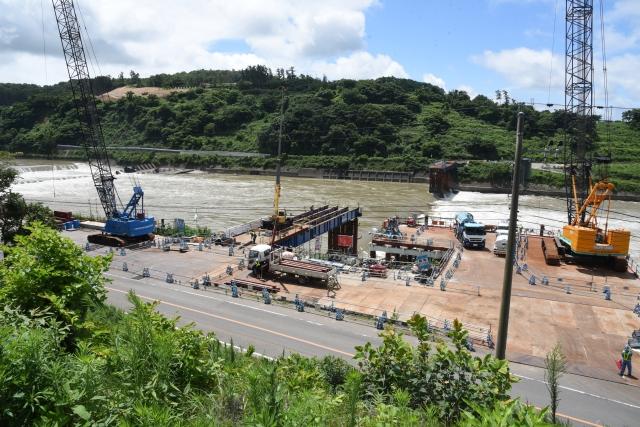 高台から見下ろす豊かな水量をたたえた川の手前側の岸辺に展開されている工事現場に、大型クレーンやトラックなど様々な建設重機が並べられている様子の写真