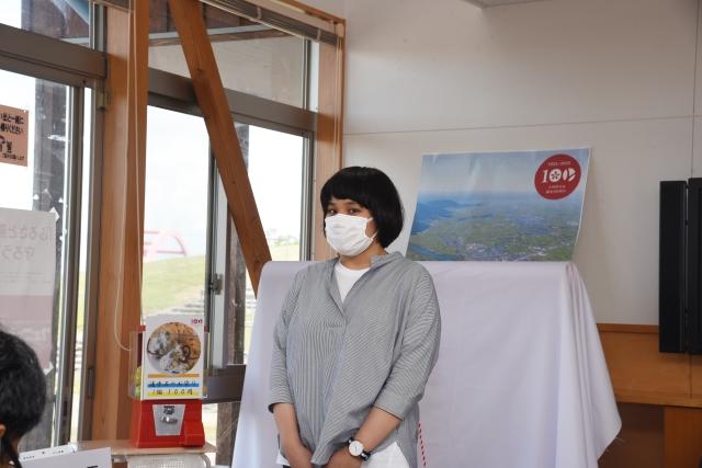 室内で序幕前の小型の神社を前に挨拶をしている、マスクを付け灰色のカットシャツを着た女性の写真