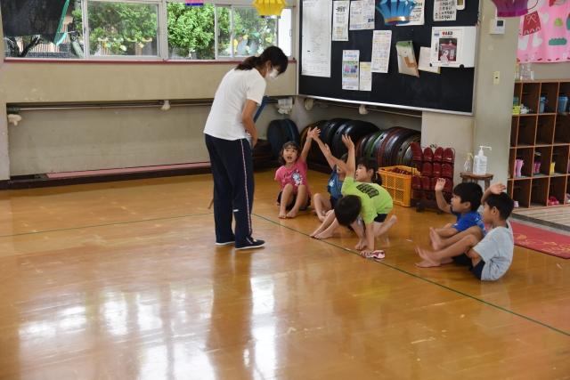 幼稚園の室内に立ちジャージ姿で呼びかけを行う先生と、その前で座りながら手を挙げている6人の子どもたちの写真