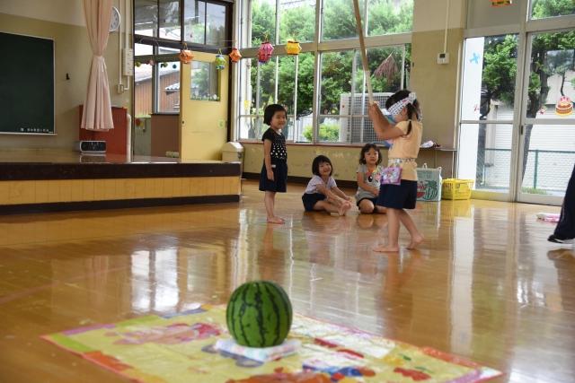 幼稚園の室内で目隠しをしてスイカ割の棒を振りかざしながらスイカを探して歩いている女の子と、その後ろで声をかけている子どもたちの写真