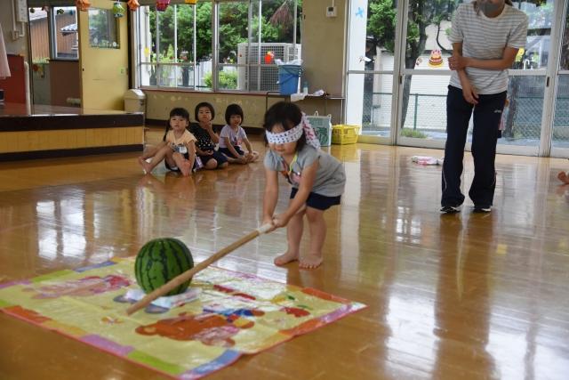幼稚園の室内に広げられたシートの上に置いてあるスイカの僅かに隣を棒でたたいてしまっている目隠しをした女の子と、それを後ろで見守っている先生の写真