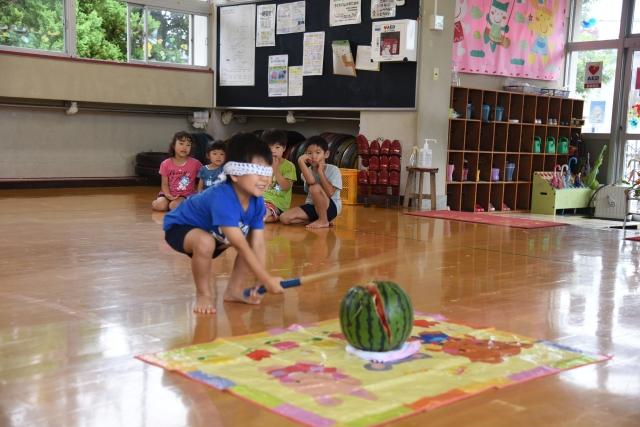 幼稚園の室内に広げられたシートの上に置いてあるスイカをめがけて屈んで棒を振り下ろし見事に叩き割る目隠しをした男の子と、それを後ろで見ている子どもたちの写真