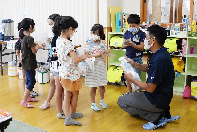 教室の中で屈みながら子供たち一人一人にマスクを手渡ししている男性職員の写真