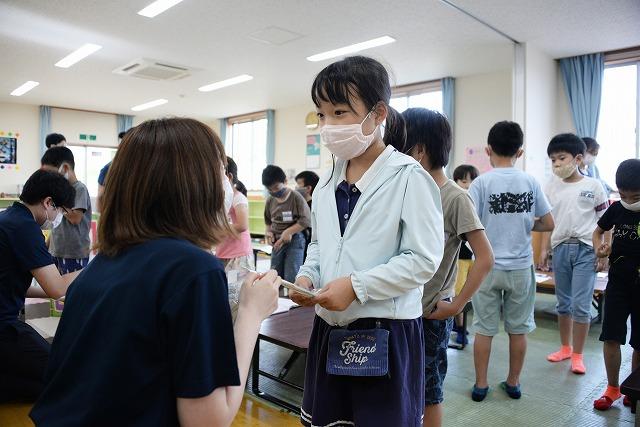 教室の中で列を作る子供達の先頭の女の子にマスクを手渡ししている女性職員の写真