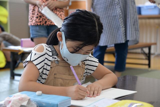 机の上にノートを開いてペンを持ち、マスクを付けて勉強をしている女の子の写真