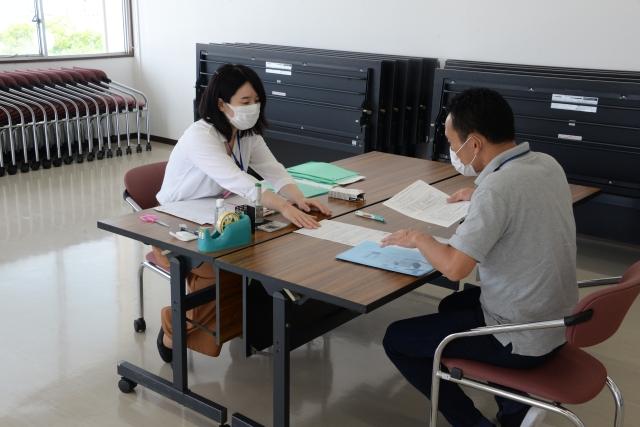 机を挟んで座り、用紙を右手に持つ男性と机の上で別の用紙を差し出している女性職員の写真
