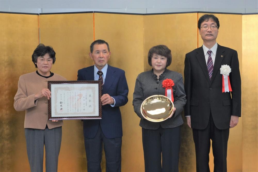 表彰状を持つ二人の男女と金色の皿のようなものを持つ女性と、その横に立つ男性の写真
