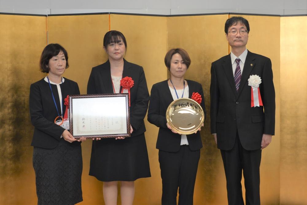 表彰状を持つ二人の女性と金色の皿のようなものを持つ女性と、その横に立つ男性の写真
