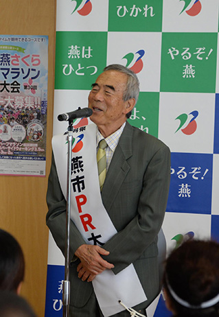 マイク前で演説する新潟県燕市PR大使のたすきをしている笑顔でスーツ姿の男性の写真