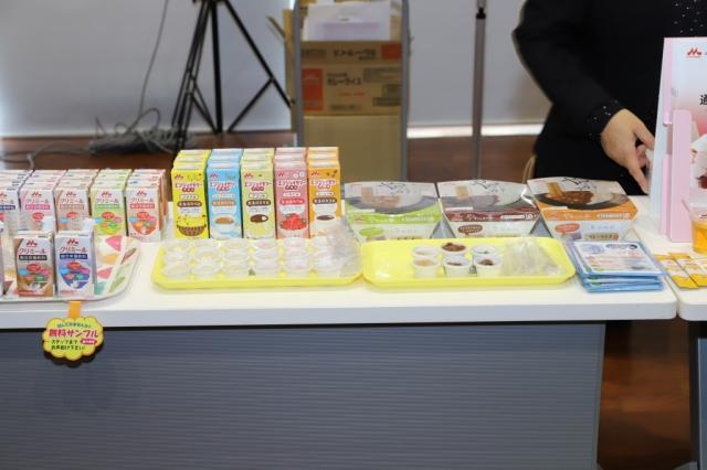 机の上に並べられた様々な介護食のパッケージサンプルとトレーの上に並べられ小皿に入った試食用の介護食の写真