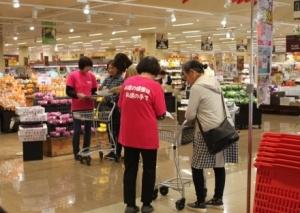 スーパーで、ピンクの服を着た人たちが、籠を持った人たちに質問をしている写真