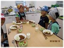カレーとサラダが並んだテーブルに向かって、三角巾をつけた子どもたちが着席している写真