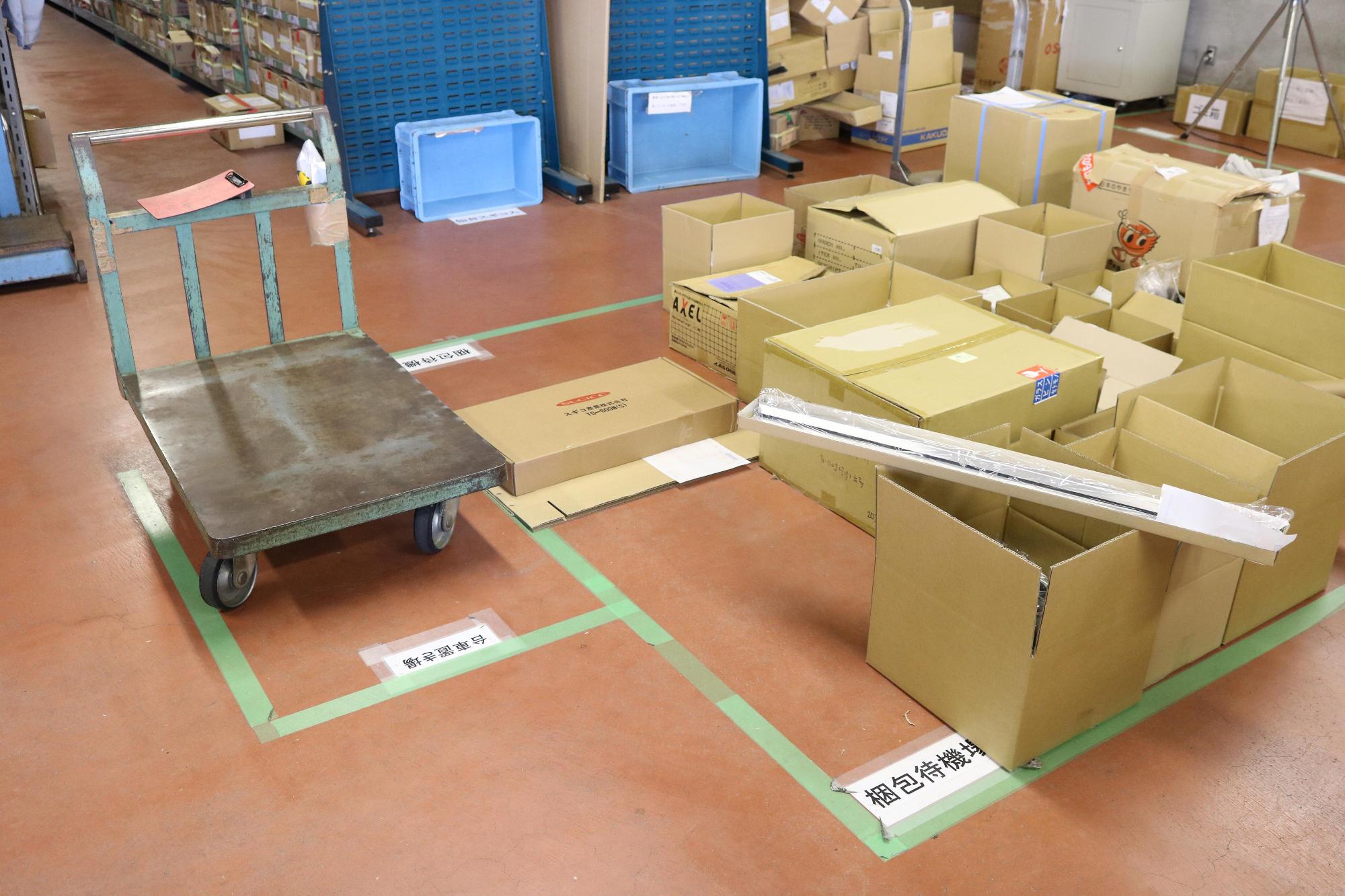緑のテープの枠に台車や箱の荷物が多数置かれている写真