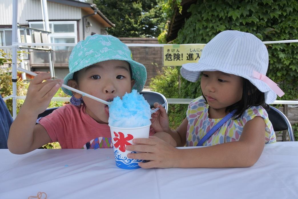 2人の女の子が水色と白の帽子を被ってテーブルに座って、水色の帽子の子がストローで水色のシロップがかかった山盛りのかき氷を食べている写真