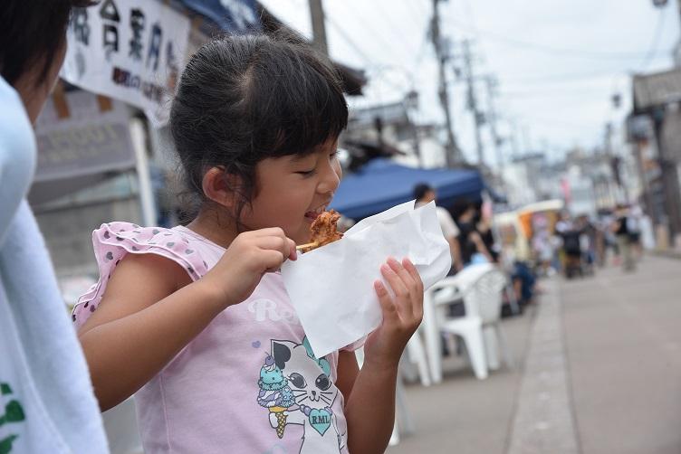 半袖のピンクの洋服を着た女の子が左手に紙ずつみ、右手に串の食べ物をもって、串ものを食べている写真