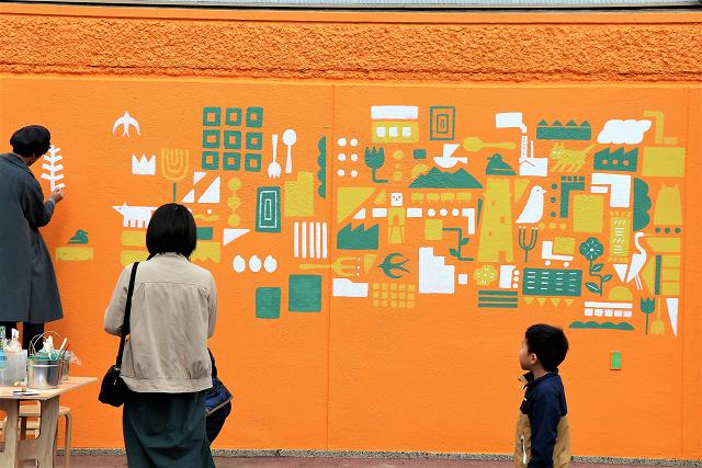 アーティストがオレンジ色の壁に絵を描いている姿を見ている親子連れの写真