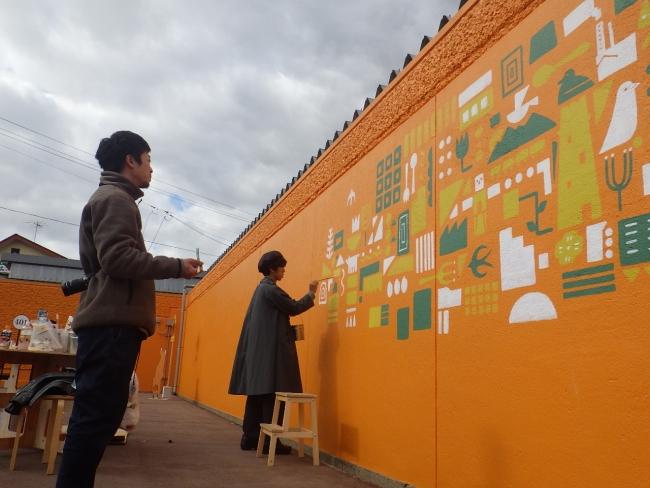 オレンジ色の庁舎壁面に、白、黄色、緑色の図形を描いていくアーティストの写真